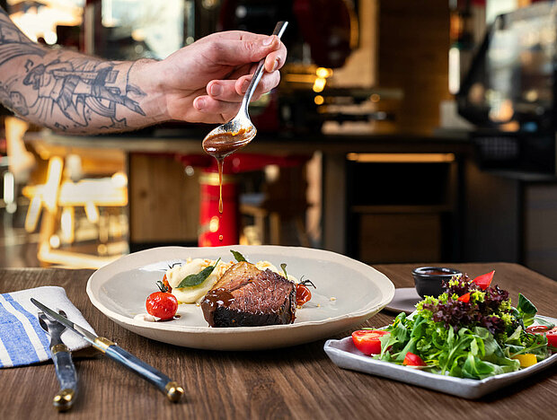 Tätowierte Hand tropft Soße auf ein Stück Fleisch mit Püree, im Hintergrund ein Beilagensalat und Restaurantdekoration