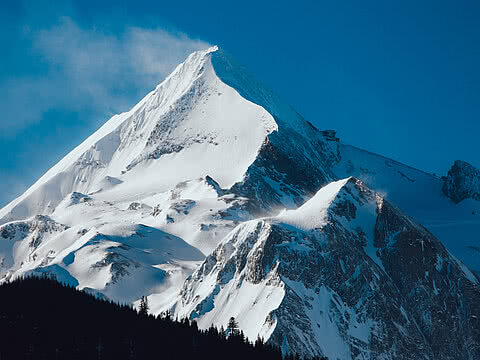 Nahaufnahme eines schneebedeckten Berggipfels unter klarem, blauem Himmel. Die weißen Hänge und felsigen Bereiche leuchten in der Sonne.