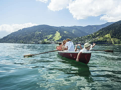 Familie sitzt in einem roten Ruderboot auf einem See, umgeben von grünen Bergen. Ein Junge mit Hut streckt sich nach dem Wasser, während die Sonne scheint.