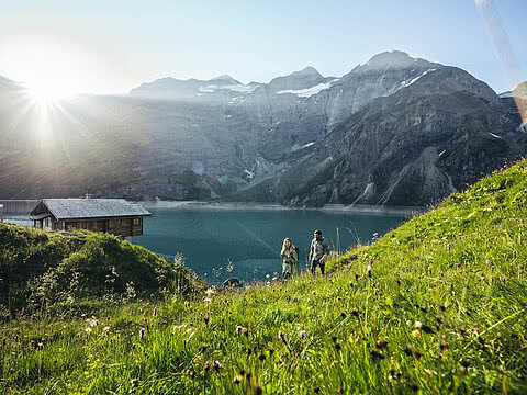 Zwei Wanderer auf einer grünen Wiese mit Blick auf einen Bergsee bei Sonnenaufgang. Umgeben von hohen Bergen und idyllischer Natur.