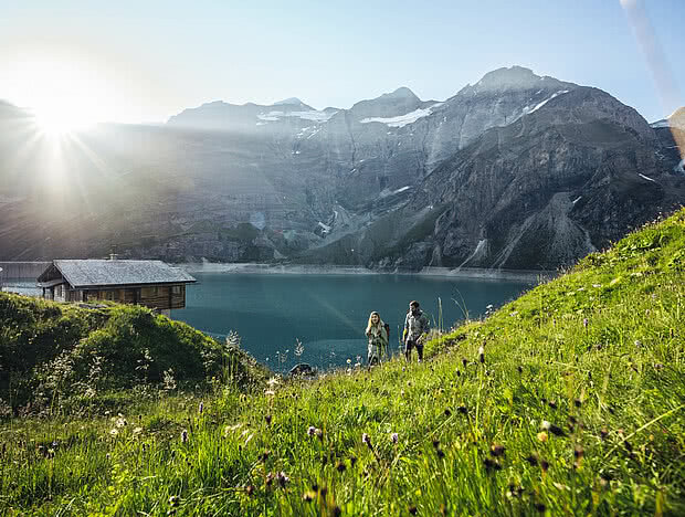Zwei Wanderer auf einer grünen Wiese mit Blick auf einen Bergsee bei Sonnenaufgang. Umgeben von hohen Bergen und idyllischer Natur.