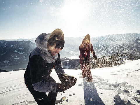 Zwei Personen bei einer Schneeballschlacht auf einem verschneiten Berghang. Sonniges Wetter und Alpenpanorama im Hintergrund.