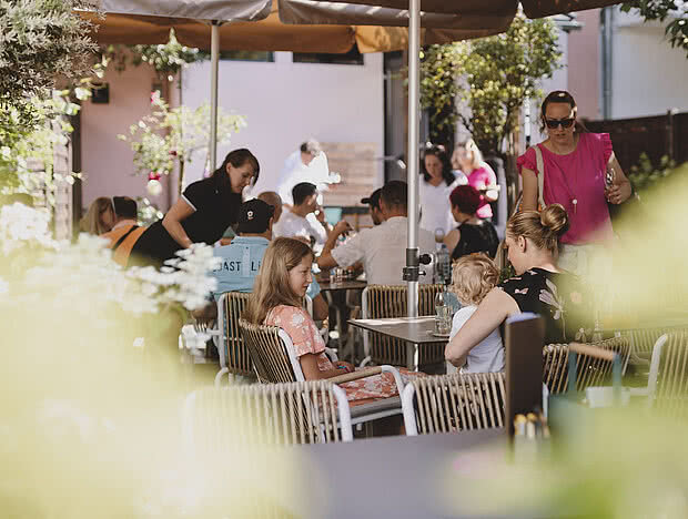 Belebter Außenbereich eines Cafés mit Gästen an mehreren Tischen, umgeben von Pflanzen und Sonnenschirmen.