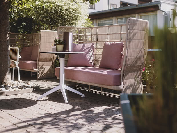 Gemütlicher Außenbereich eines Cafés mit rosa gepolsterten Sofas, weißen Tischen und Stühlen, umgeben von grünen Pflanzen und natürlichem Schatten.