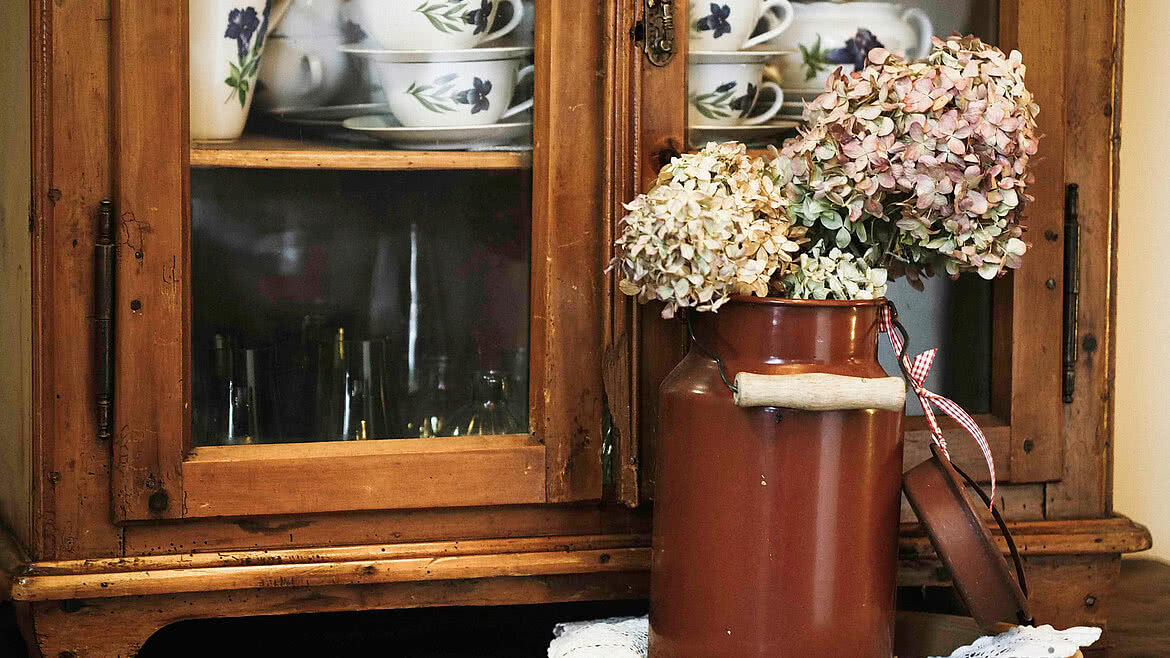 Detailaufnahme eines rustikalen Holzschranks mit Porzellangeschirr, dekoriert mit getrockneten Hortensien in einem nostalgischen braunen Milchkrug, auf einer weißen Spitzendecke.