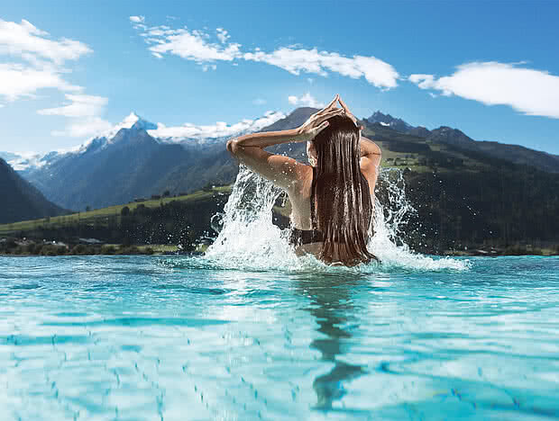 Frau in einem Pool mit Blick auf ein beeindruckendes Bergpanorama. Sie trägt lange Haare und hebt die Arme, während Wasser spritzt. Sommerliche Atmosphäre unter klarem Himmel.