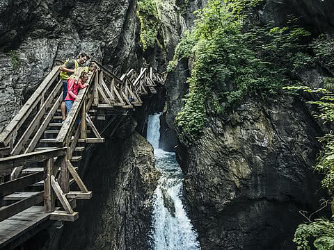 Familie auf einem hölzernen Steg in einer beeindruckenden Schlucht mit Wasserfall. Steile Felswände und grüne Vegetation umrahmen die Szene.