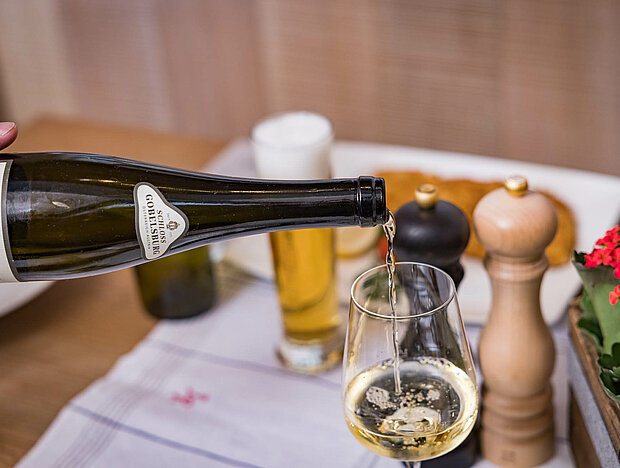 Eine Hand gießt Weißwein aus einer Flasche in ein Weinglas, im Hintergrund Salz- und Pfefferstreuer