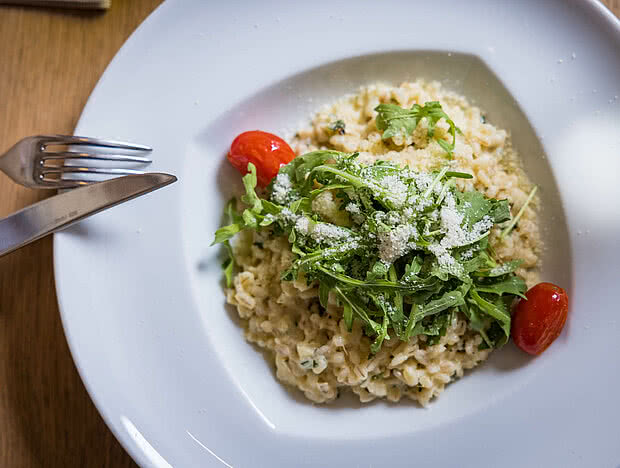 Ein Teller mit cremigem Risotto, garniert mit frischem Rucola und zwei Kirschtomaten, auf einem gedeckten Tisch