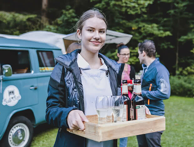 Junge Servicekraft hält ein Holztablett mit Getränken, darunter Sektgläser und Bierflaschen. Im Hintergrund ein Retro-Foodtruck und eine grüne Wiese.