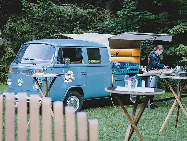 Blauer Volkswagen Bulli mit geöffneter Klappe als mobile Bar bei einer Outdoor-Veranstaltung. Eine Frau arrangiert Speisen, während Stehtische das Setup ergänzen.