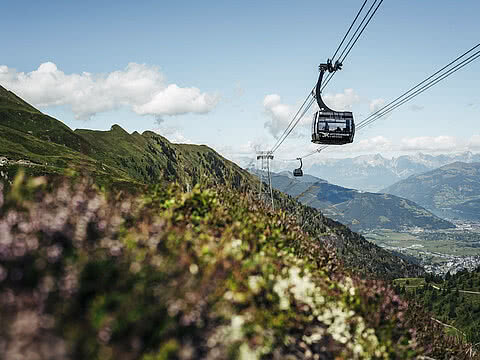 Moderne Seilbahn schwebt über einer grünen Berglandschaft. Im Hintergrund das Panorama von Tälern und Bergketten unter blauem Himmel.