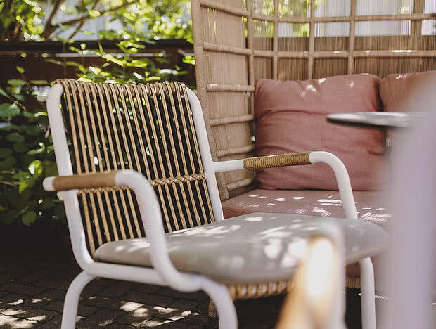 Detailaufnahme eines weißen Rattanstuhls in einem Café-Außenbereich, umgeben von grünem Laub und schattigen Plätzen.