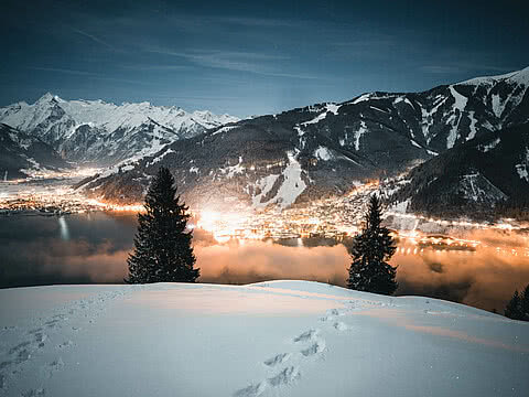 Zell am See bei Nacht, beleuchtet mit Blick auf schneebedeckte Berge und den glitzernden See. Spuren im Schnee im Vordergrund.