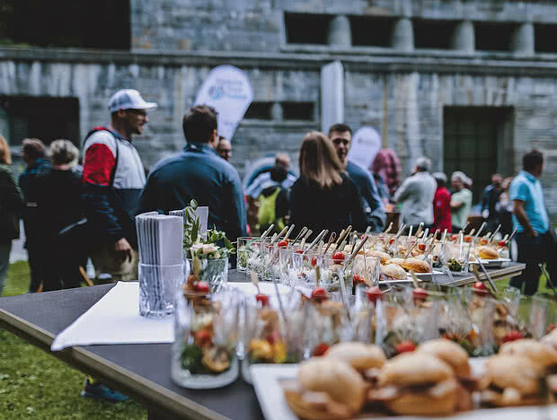 Outdoor-Event mit Gästen und einer Auswahl an Fingerfood auf einem Tisch, darunter Gläser mit Tomaten und gegrilltem Gemüse sowie belegte Brötchen. Im Hintergrund sind Gäste in Gespräch vertieft.