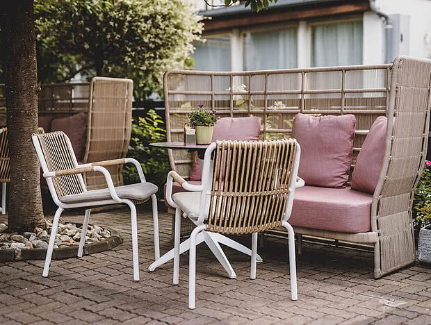 Außenbereich eines Cafés mit einer gemütlichen Sitzgruppe aus weißen Stühlen und rosa gepolsterten Sofas, schattiert von Bäumen und umgeben von Pflanzen.