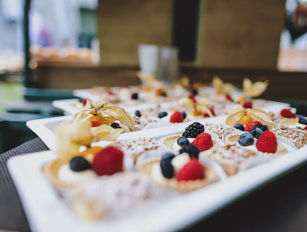 Nahaufnahme von Dessert-Törtchen mit frischen Beeren wie Himbeeren, Brombeeren und Physalis, serviert auf einem weißen Tablett bei einem Event im Freien.